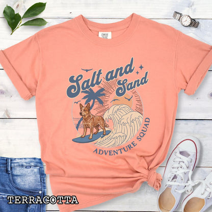Orange Comfort Colors t-shirt for Australian Cattle Dog lovers with beach-themed graphic of a surfing Red Heeler, waves and sunset, and the text 'Salt and Sand Adventure Squad' in retro lettering, shown on a wooden surface with accessories.