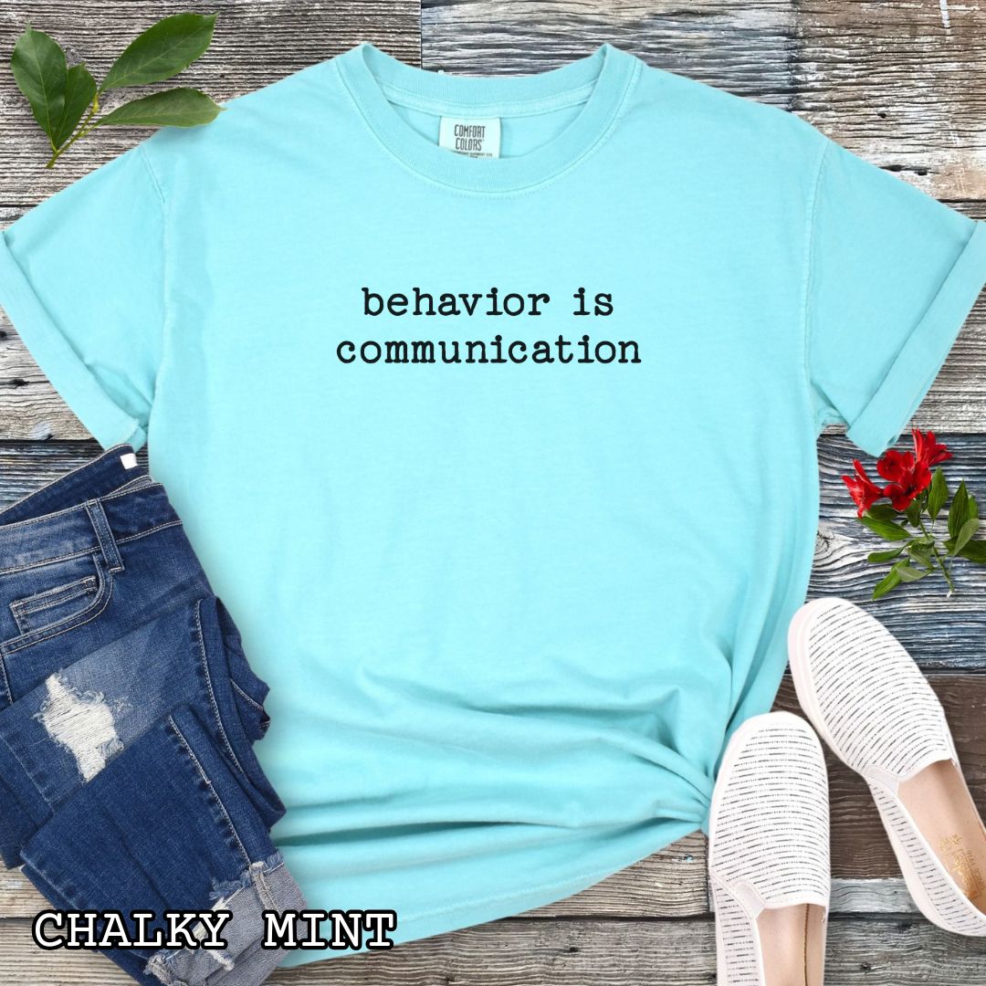 Mint Comfort Colors t-shirt for reactive dog trainers and behavior-focused trainers, with 'behavior is communication' text in typewriter font, shown on a wooden surface with sunglasses, jeans, and flowers.