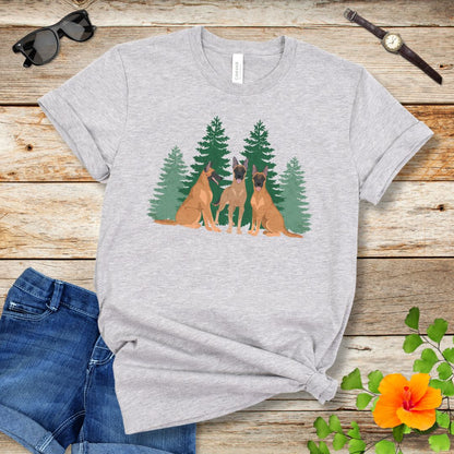 A light grey unisex t-shirt with an image of 3 Belgian Malinois dogs and evergreen trees printed on it, displayed flat with a pair of jeans and white shoes on a wooden surface.