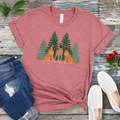A mauve unisex t-shirt with an image of 3 Belgian Malinois dogs and evergreen trees printed on it, displayed flat with a pair of jeans and white shoes on a wooden surface.
