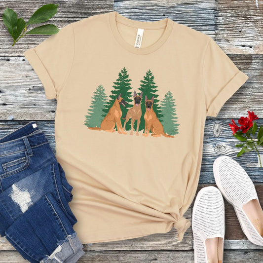 A cream-colored unisex t-shirt with an image of 3 Belgian Malinois dogs and evergreen trees printed on it, displayed flat with a pair of jeans and white shoes on a wooden surface.