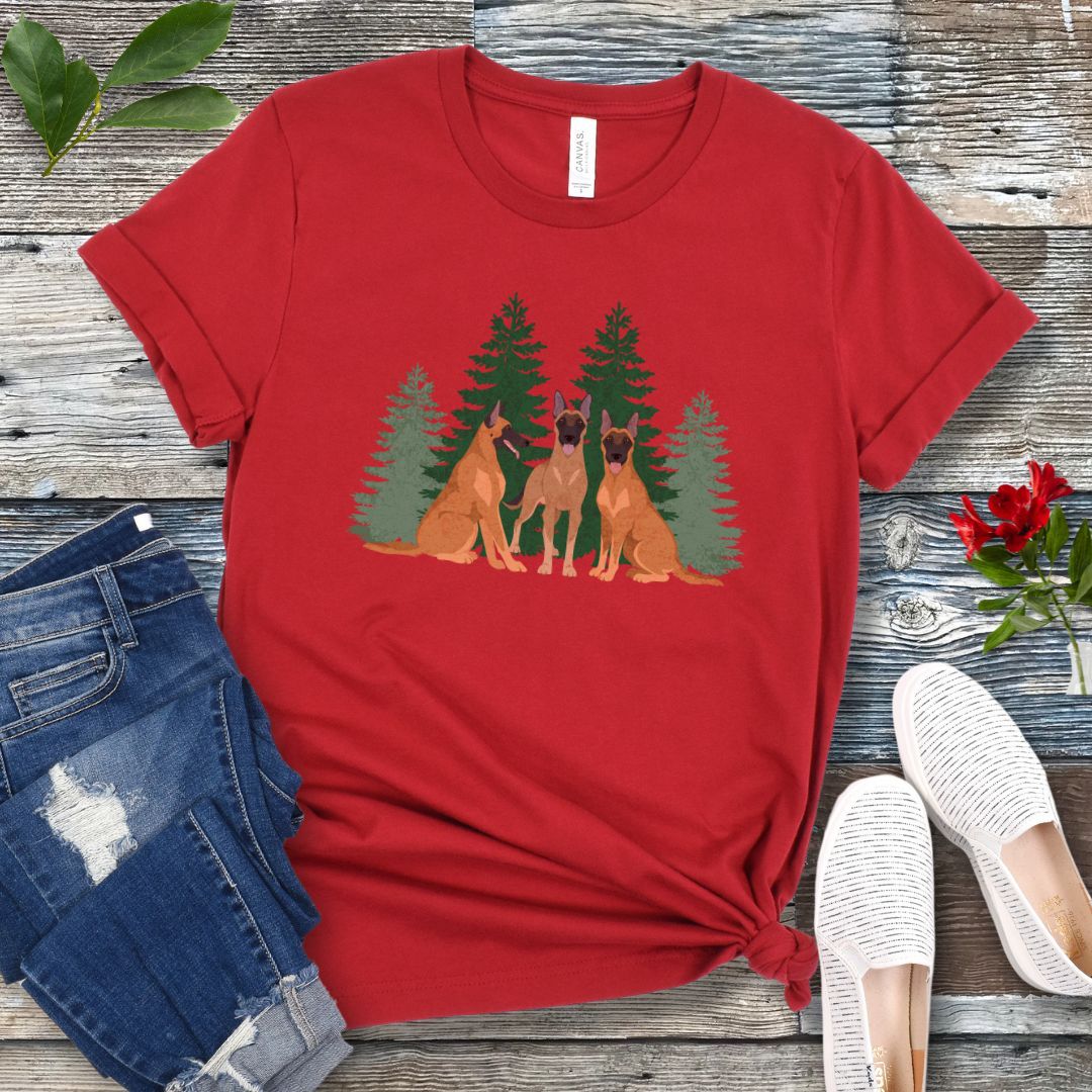 A dark red unisex t-shirt with an image of 3 Belgian Malinois dogs and evergreen trees printed on it, displayed flat with a pair of jeans and white shoes on a wooden surface.