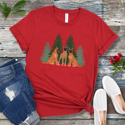 A dark red unisex t-shirt with an image of 3 Belgian Malinois dogs and evergreen trees printed on it, displayed flat with a pair of jeans and white shoes on a wooden surface.