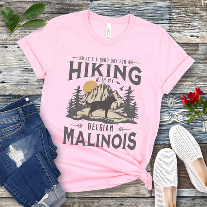 A pink t-shirt with a vintage style graphic print of a mountain range, pine trees, and a Malinois dog silhouette, with the text 'It's a good day for HIKING with my BELGIAN MALINOIS'. Presented with blue jeans and white shoes.