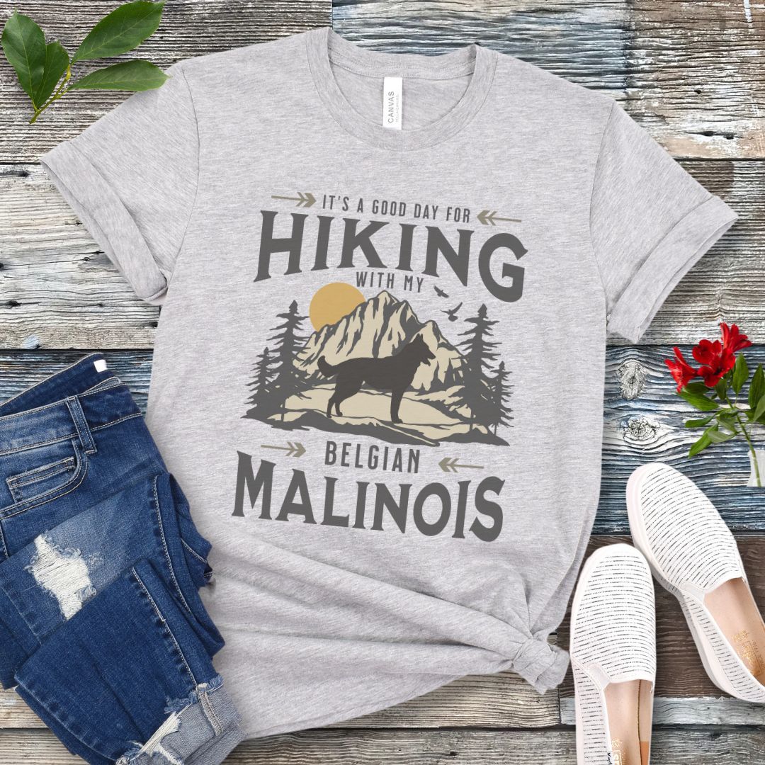 A grey t-shirt with a vintage style graphic print of a mountain range, pine trees, and a Malinois dog silhouette, with the text 'It's a good day for HIKING with my BELGIAN MALINOIS'. Presented with blue jeans and white shoes.