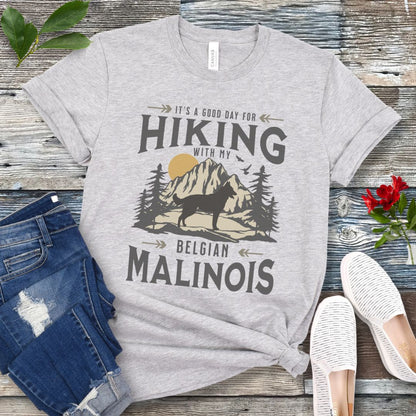 A grey t-shirt with a vintage style graphic print of a mountain range, pine trees, and a Malinois dog silhouette, with the text 'It's a good day for HIKING with my BELGIAN MALINOIS'. Presented with blue jeans and white shoes.