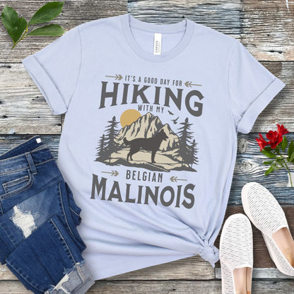 A light blue t-shirt with a vintage style graphic print of a mountain range, pine trees, and a Malinois dog silhouette, with the text 'It's a good day for HIKING with my BELGIAN MALINOIS'. Presented with blue jeans and white shoes.
