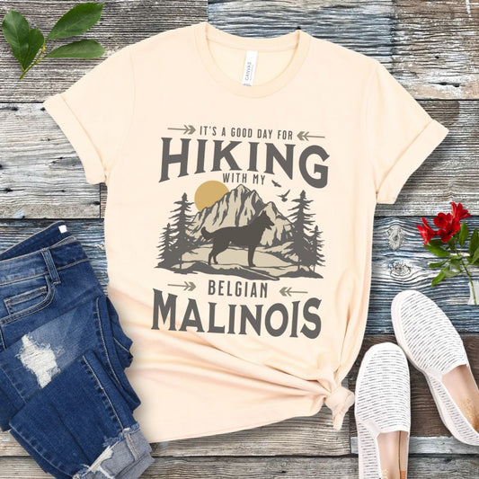 A beige colored t-shirt with a vintage style graphic print of a mountain range, pine trees, and a Malinois dog silhouette, with the text 'It's a good day for HIKING with my BELGIAN MALINOIS'. There are also images of blue jeans and white shoes next to it for scale. 
