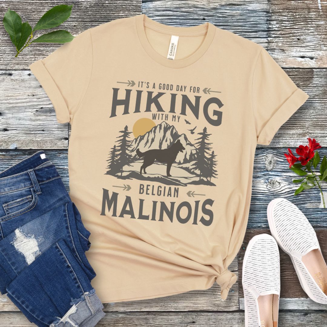 A cream-colored t-shirt with a vintage style graphic print of a mountain range, pine trees, and a Malinois dog silhouette, with the text 'It's a good day for HIKING with my BELGIAN MALINOIS'. Presented with blue jeans and white shoes.