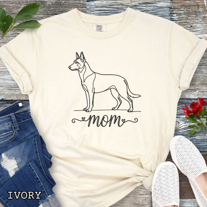 Beige Comfort Colors t-shirt with a line art Belgian Malinois dog illustration and 'mom' text in feminine script beneath, shown on a wooden surface with denim jeans and a flower.