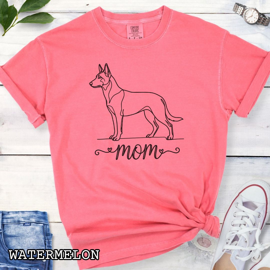 Pink Comfort Colors t-shirt with a line art Belgian Malinois dog illustration and 'mom' text in feminine script beneath, shown on a wooden surface with denim jeans and a flower.
