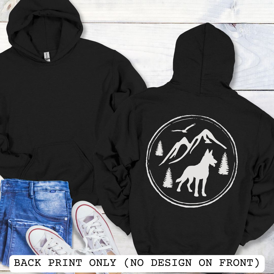 Two black Malinois hoodies shown on a white wood background, with the front view on the left and back view on the right. The back has a large white circular graphic featuring a wolf, mountains, trees, and birds.