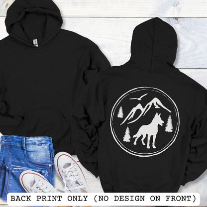 Two black Malinois hoodies shown on a white wood background, with the front view on the left and back view on the right. The back has a large white circular graphic featuring a wolf, mountains, trees, and birds.
