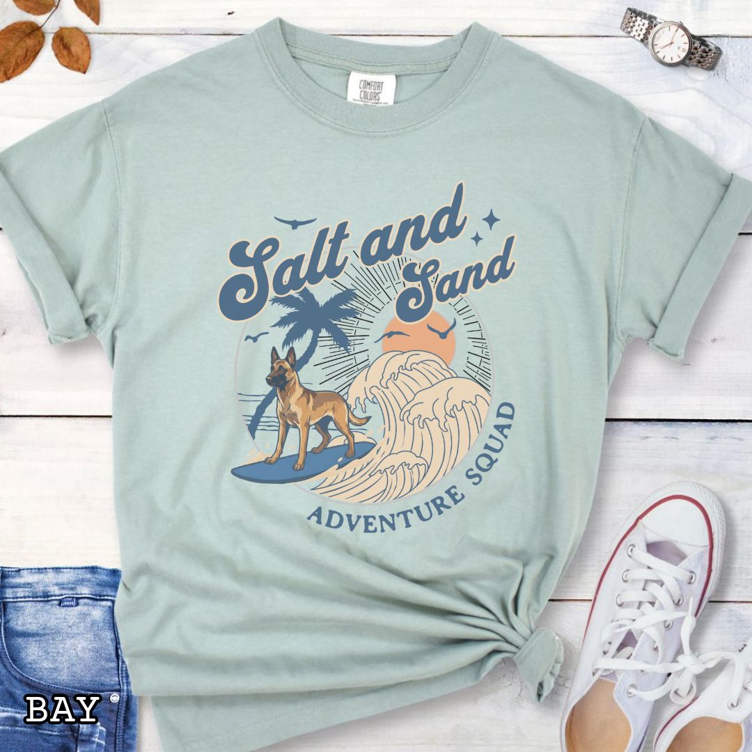 Pale green Comfort Colors t-shirt for Belgian Malinois lovers with beach-themed graphic of a surfing Malinois dog, waves and sunset, and the text 'Salt and Sand Adventure Squad' in retro lettering, shown on a wooden surface with accessories.