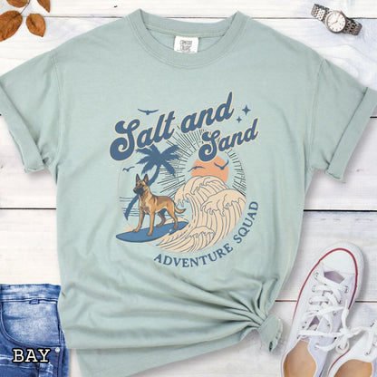 Pale green Comfort Colors t-shirt for Belgian Malinois lovers with beach-themed graphic of a surfing Malinois dog, waves and sunset, and the text 'Salt and Sand Adventure Squad' in retro lettering, shown on a wooden surface with accessories.