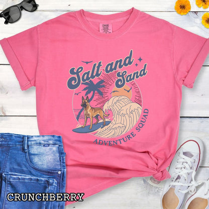 Pink Comfort Colors t-shirt for Belgian Malinois lovers with beach-themed graphic of a surfing Malinois dog, waves and sunset, and the text 'Salt and Sand Adventure Squad' in retro lettering, shown on a wooden surface with accessories.