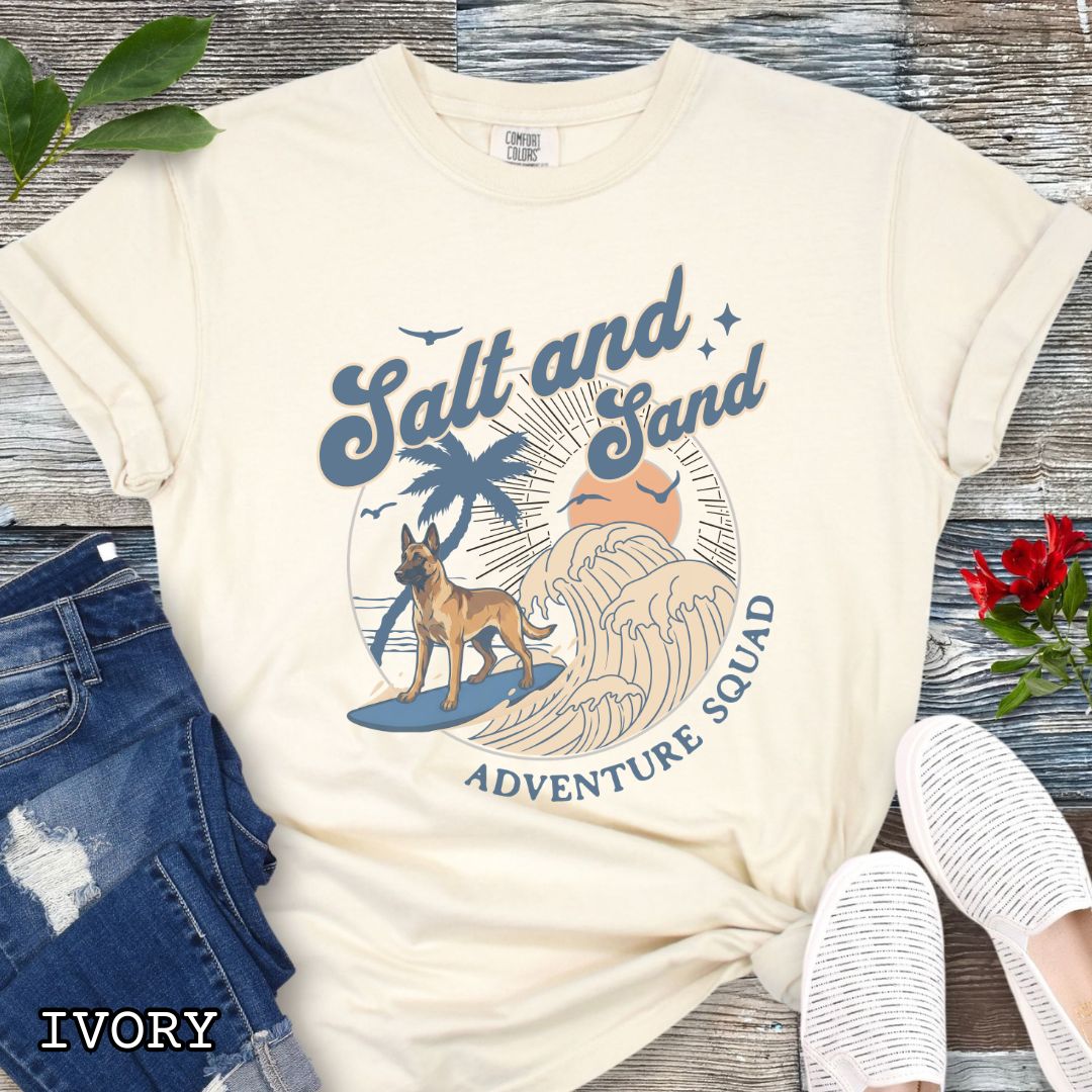 Beige Comfort Colors t-shirt for Belgian Malinois lovers with beach-themed graphic of a surfing Malinois dog, waves and sunset, and the text 'Salt and Sand Adventure Squad' in retro lettering, shown on a wooden surface with accessories.