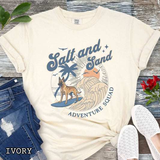Beige Comfort Colors t-shirt for Belgian Malinois lovers with beach-themed graphic of a surfing Malinois dog, waves and sunset, and the text 'Salt and Sand Adventure Squad' in retro lettering, shown on a wooden surface with accessories.