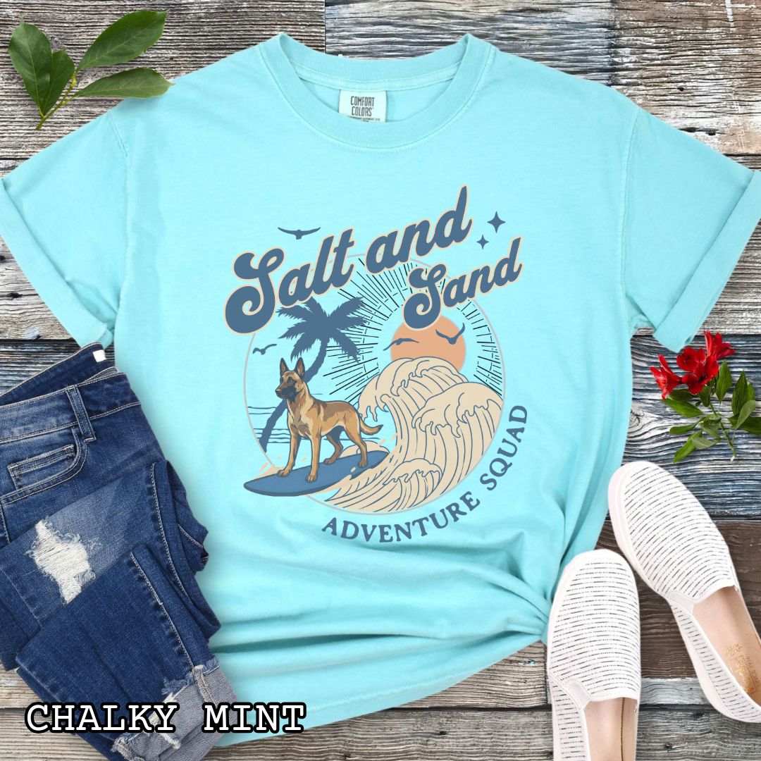 Mint Comfort Colors t-shirt for Belgian Malinois lovers with beach-themed graphic of a surfing Malinois dog, waves and sunset, and the text 'Salt and Sand Adventure Squad' in retro lettering, shown on a wooden surface with accessories.
