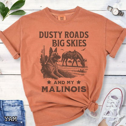 Yam-orange Comfort Colors t-shirt featuring a Western-style design with grazing horse, rustic fence, and a Malinois illustration; text reads “Dusty Roads, Big Skies, and My Malinois.”
