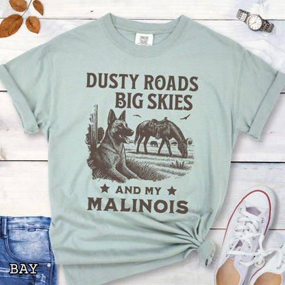 Light green Comfort Colors t-shirt featuring a Western-style design with grazing horse, rustic fence, and a Malinois illustration; text reads “Dusty Roads, Big Skies, and My Malinois.”