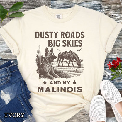 Ivory Comfort Colors t-shirt featuring a Western-style design with grazing horse, rustic fence, and a Malinois illustration; text reads “Dusty Roads, Big Skies, and My Malinois.”