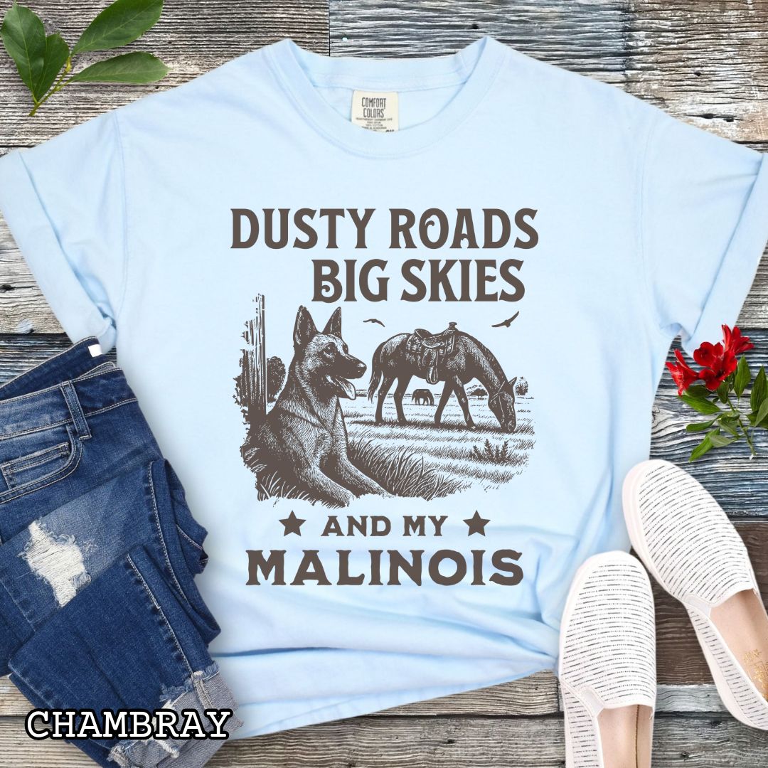 Light blue Comfort Colors t-shirt featuring a Western-style design with grazing horse, rustic fence, and a Malinois illustration; text reads “Dusty Roads, Big Skies, and My Malinois.”