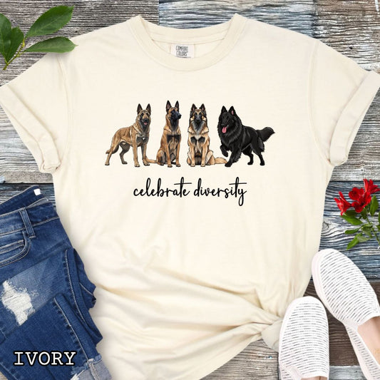 Beige Comfort Colors t-shirt with Belgian Laekenois, Sheepdog, Tervuren and Malinois dog illustrations and 'celebrate diversity' text on a wooden surface.