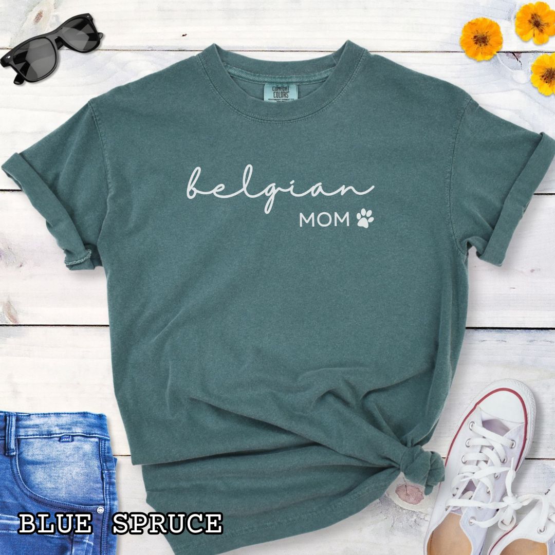 Green Comfort Colors t-shirt for Belgian Sheepdog lovers with 'belgian mom' text design in feminine handwritten script, accompanied by a small paw print, shown on a wooden surface with sunglasses, jeans, and sneakers.