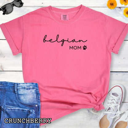Pink Comfort Colors t-shirt for Belgian Sheepdog lovers with 'belgian mom' text design in feminine handwritten script, accompanied by a small paw print, shown on a wooden surface with sunglasses, jeans, and sneakers.