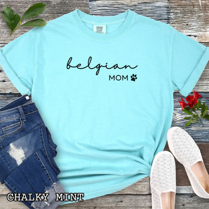 Mint Comfort Colors t-shirt for Belgian Sheepdog lovers with 'belgian mom' text design in feminine handwritten script, accompanied by a small paw print, shown on a wooden surface with sunglasses, jeans, and sneakers.