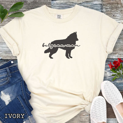 Beige t-shirt with Belgian Sheepdog silhouette and 'Belgian Mom' text in handwritten script lettering, shown on a wooden surface with denim jeans and a flower.