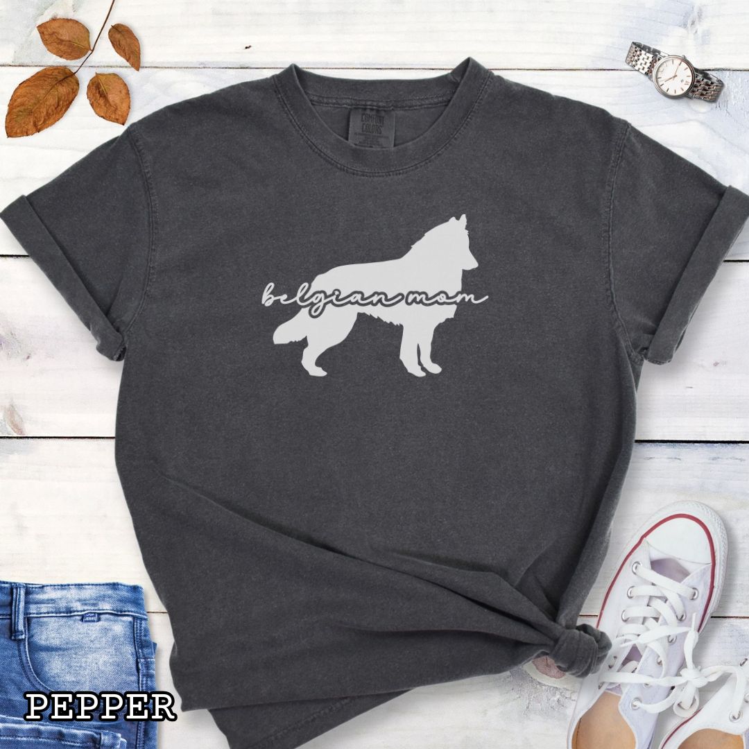 Dark grey t-shirt with Belgian Sheepdog silhouette and 'Belgian Mom' text in handwritten script lettering, shown on a wooden surface with denim jeans and a flower.