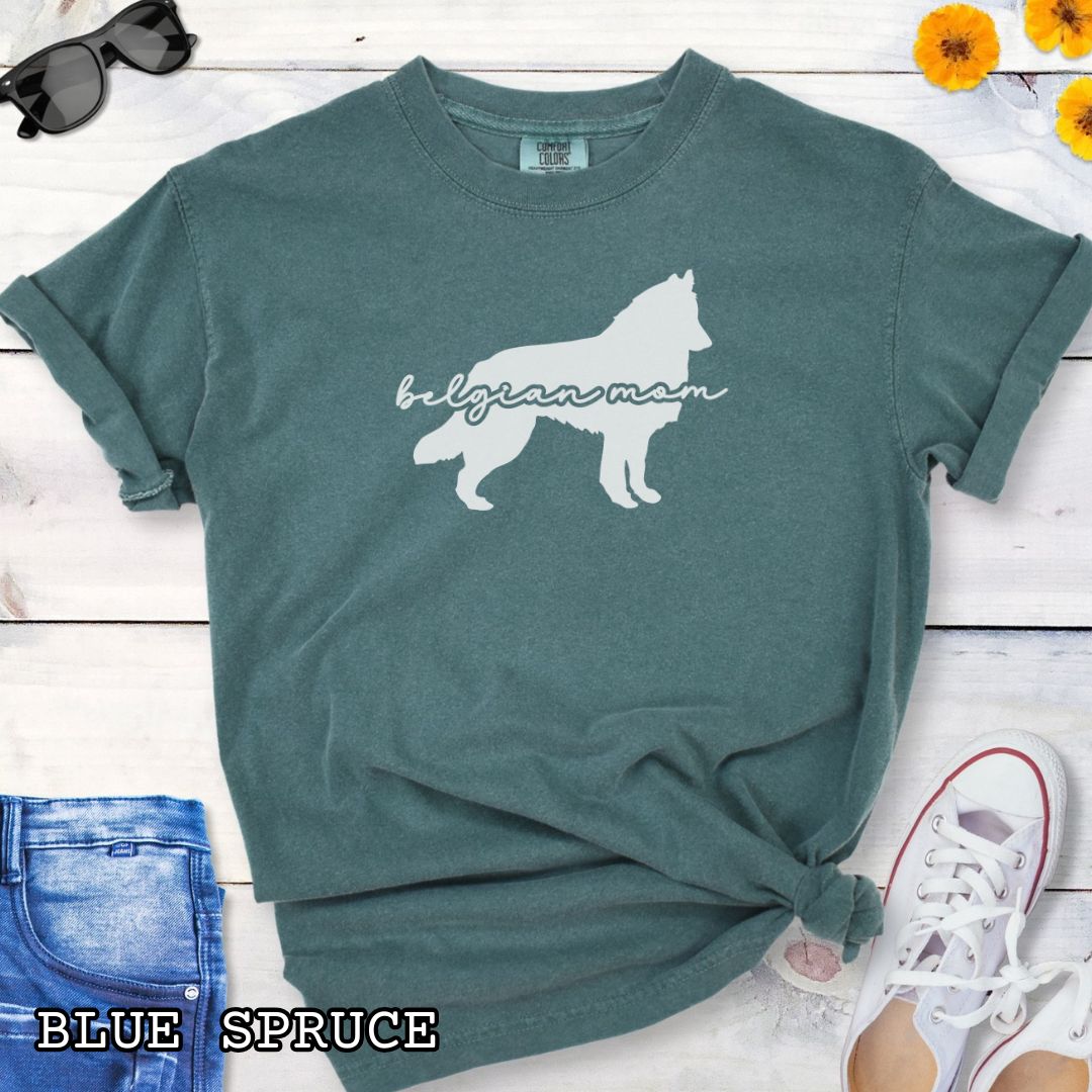 Green t-shirt with Belgian Sheepdog silhouette and 'Belgian Mom' text in handwritten script lettering, shown on a wooden surface with denim jeans and a flower.