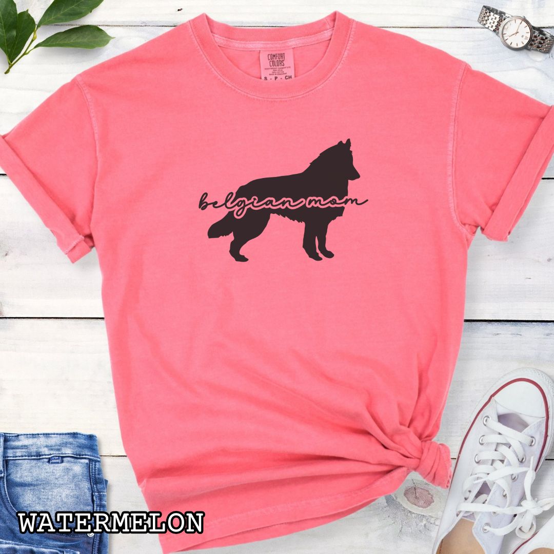 Pink t-shirt with Belgian Sheepdog silhouette and 'Belgian Mom' text in handwritten script lettering, shown on a wooden surface with denim jeans and a flower.