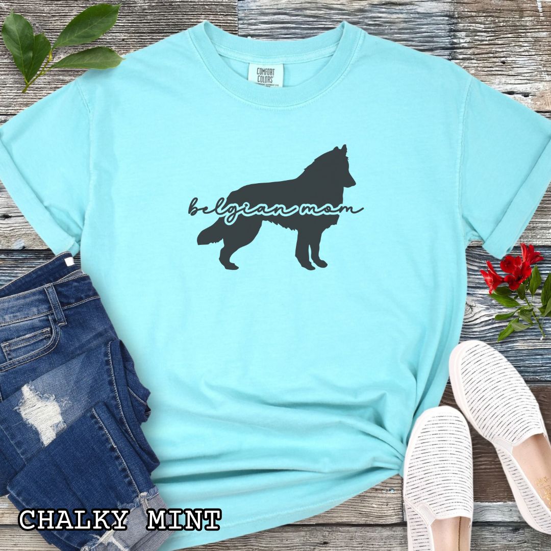 Teal t-shirt with Belgian Sheepdog silhouette and 'Belgian Mom' text in handwritten script lettering, shown on a wooden surface with denim jeans and a flower.