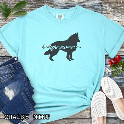 Teal t-shirt with Belgian Sheepdog silhouette and 'Belgian Mom' text in handwritten script lettering, shown on a wooden surface with denim jeans and a flower.