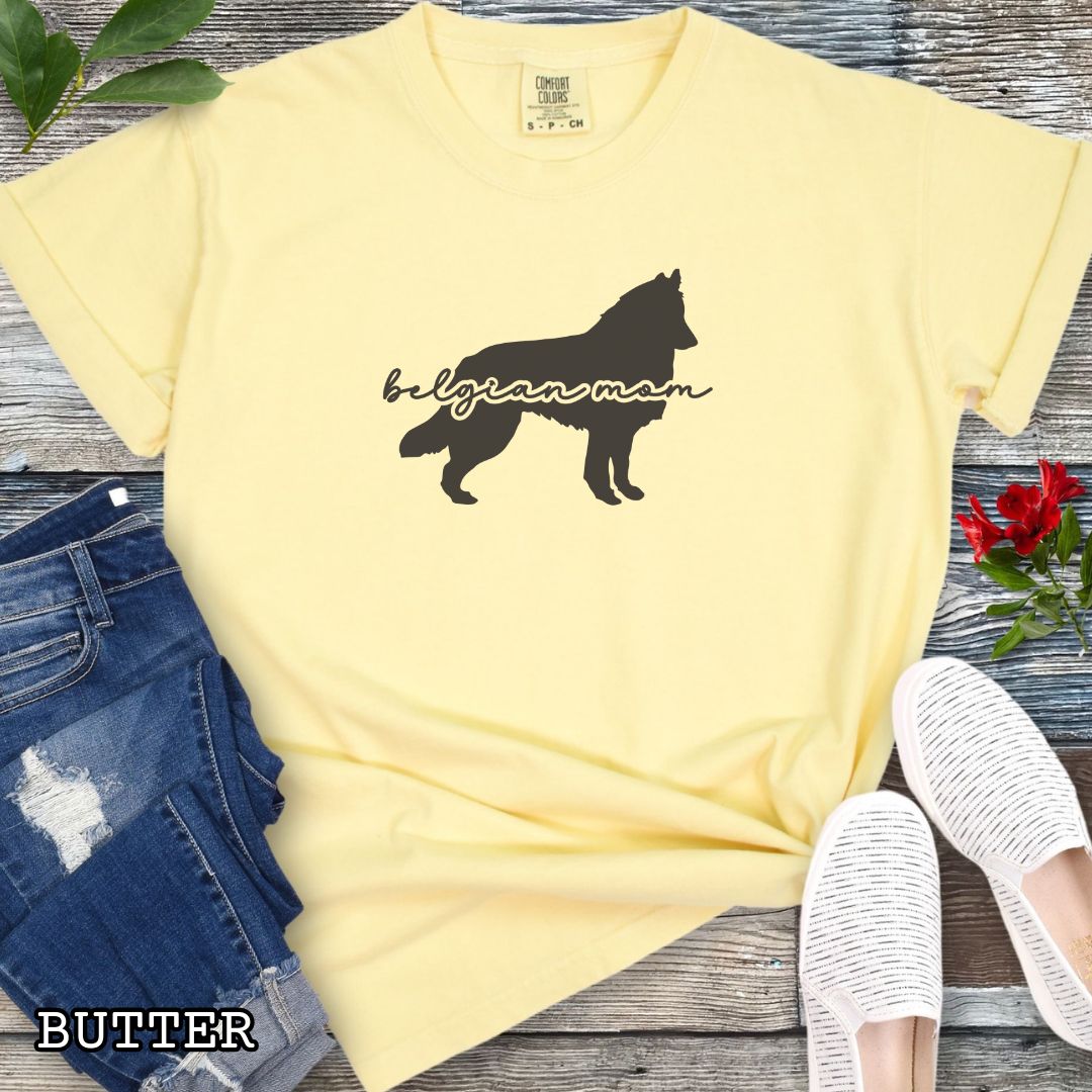 Yellow t-shirt with Belgian Sheepdog silhouette and 'Belgian Mom' text in handwritten script lettering, shown on a wooden surface with denim jeans and a flower.