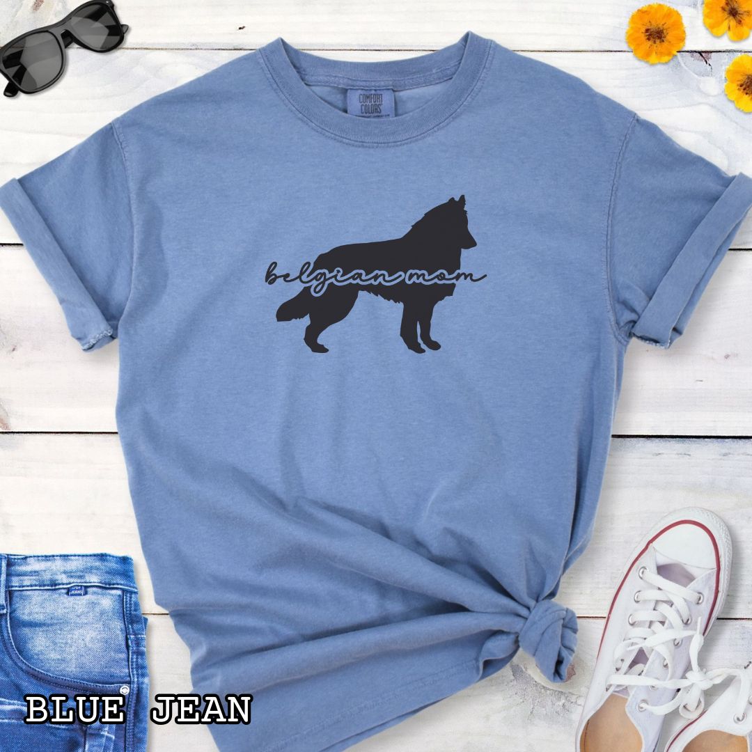 Blue t-shirt with Belgian Sheepdog silhouette and 'Belgian Mom' text in handwritten script lettering, shown on a wooden surface with denim jeans and a flower.