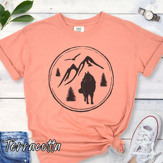 Terracotta Comfort Colors t-shirt for outdoorsy Belgian Shepherd lovers, with mountain scene and Belgian Sheepdog/Tervuren graphic, shown on a wooden surface with jeans and sneakers.