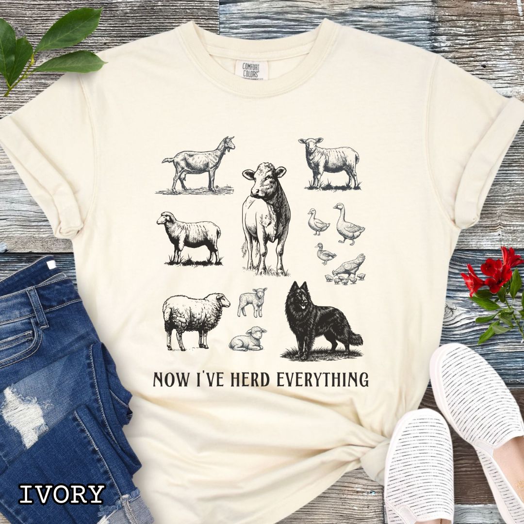 Beige Comfort Colors t-shirt for Belgian Sheepdog lovers with vintage  illustrations of farm animals and a Groenendael dog, with the humorous text 'Now I've Herd Everything', shown on a wooden surface with denim jeans and flowers.