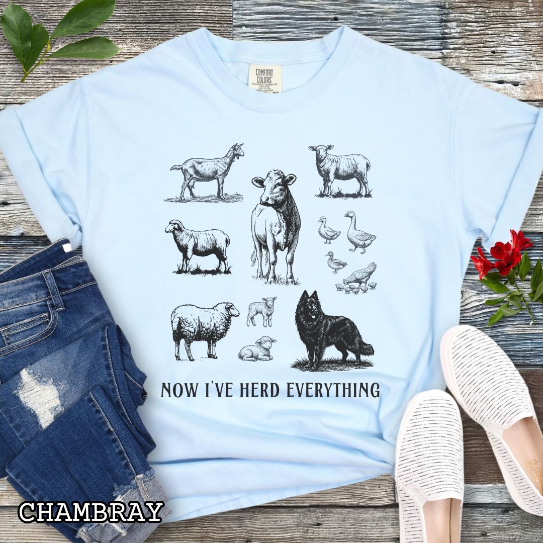 Light blue Comfort Colors t-shirt for Belgian Sheepdog lovers with vintage  illustrations of farm animals and a Groenendael dog, with the humorous text 'Now I've Herd Everything', shown on a wooden surface with denim jeans and flowers.