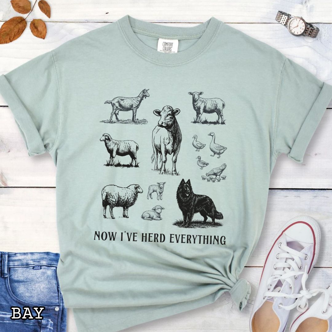 Light green Comfort Colors t-shirt for Belgian Sheepdog lovers with vintage  illustrations of farm animals and a Groenendael dog, with the humorous text 'Now I've Herd Everything', shown on a wooden surface with denim jeans and flowers.