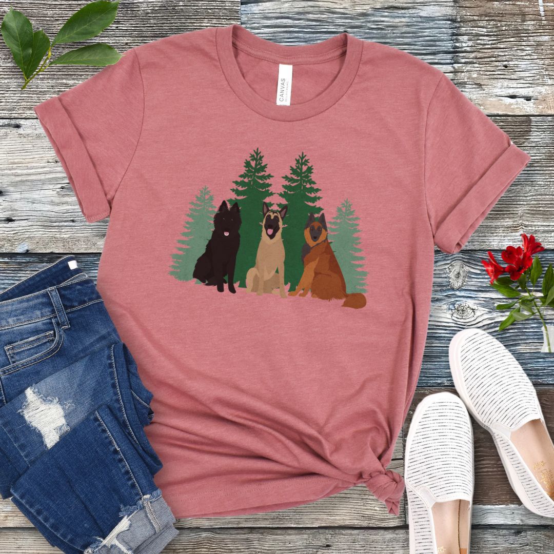 A mauve Bella Canvas 3001 unisex short sleeve t-shirt with an image of three Belgian Shepherds (Belgian Sheepdog, Malinois and Tervuren) and evergreen trees on the front, displayed on a wooden surface with a pair of jeans and white shoes in the background.