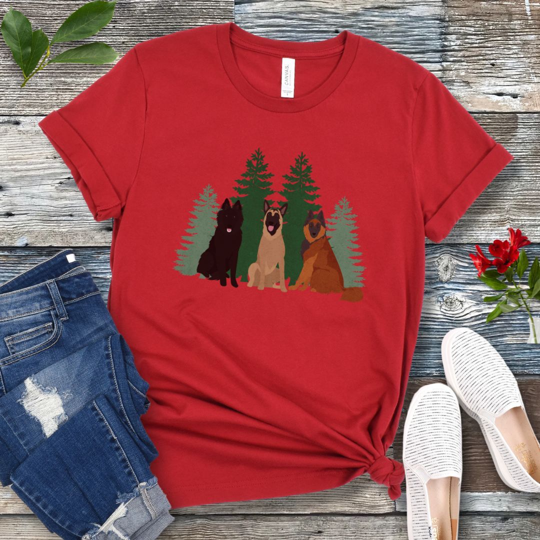 A red Bella Canvas 3001 unisex short sleeve t-shirt with an image of three Belgian Shepherds (Belgian Sheepdog, Malinois and Tervuren) and evergreen trees on the front, displayed on a wooden surface with a pair of jeans and white shoes in the background.