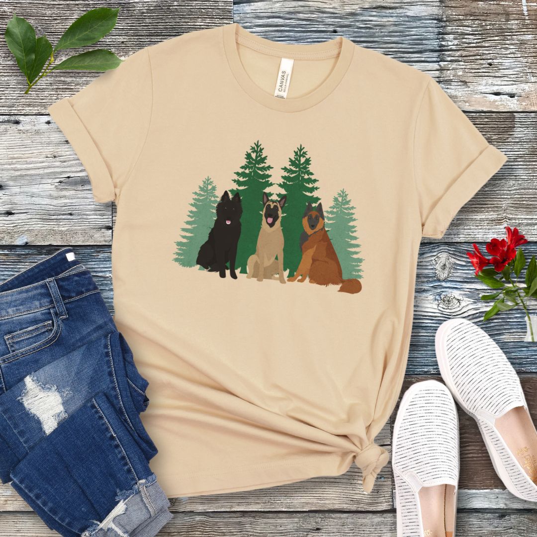 A cream Bella Canvas 3001 unisex short sleeve t-shirt with an image of three Belgian Shepherds (Belgian Sheepdog, Malinois and Tervuren) and evergreen trees on the front, displayed on a wooden surface with a pair of jeans and white shoes in the background.