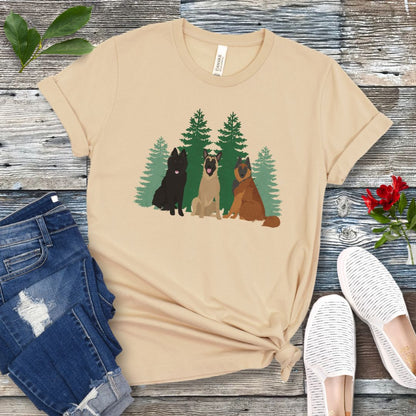 A cream Bella Canvas 3001 unisex short sleeve t-shirt with an image of three Belgian Shepherds (Belgian Sheepdog, Malinois and Tervuren) and evergreen trees on the front, displayed on a wooden surface with a pair of jeans and white shoes in the background.
