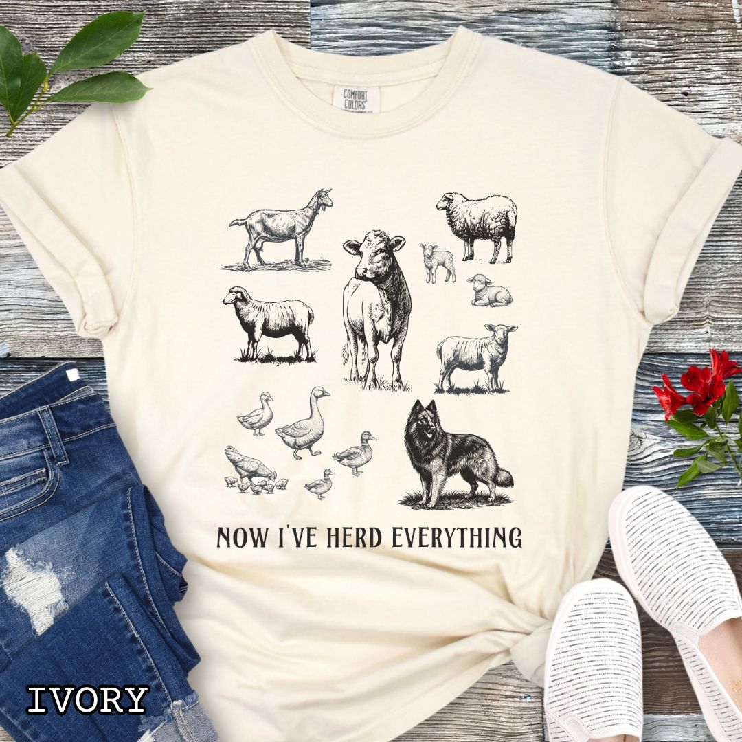 Beige Comfort Colors t-shirt for Belgian Tervuren lovers with vintage farm animal illustrations and a Belgian Tervuren dog, with the humorous text 'Now I've Herd Everything', shown on a wooden surface with denim jeans and flowers.