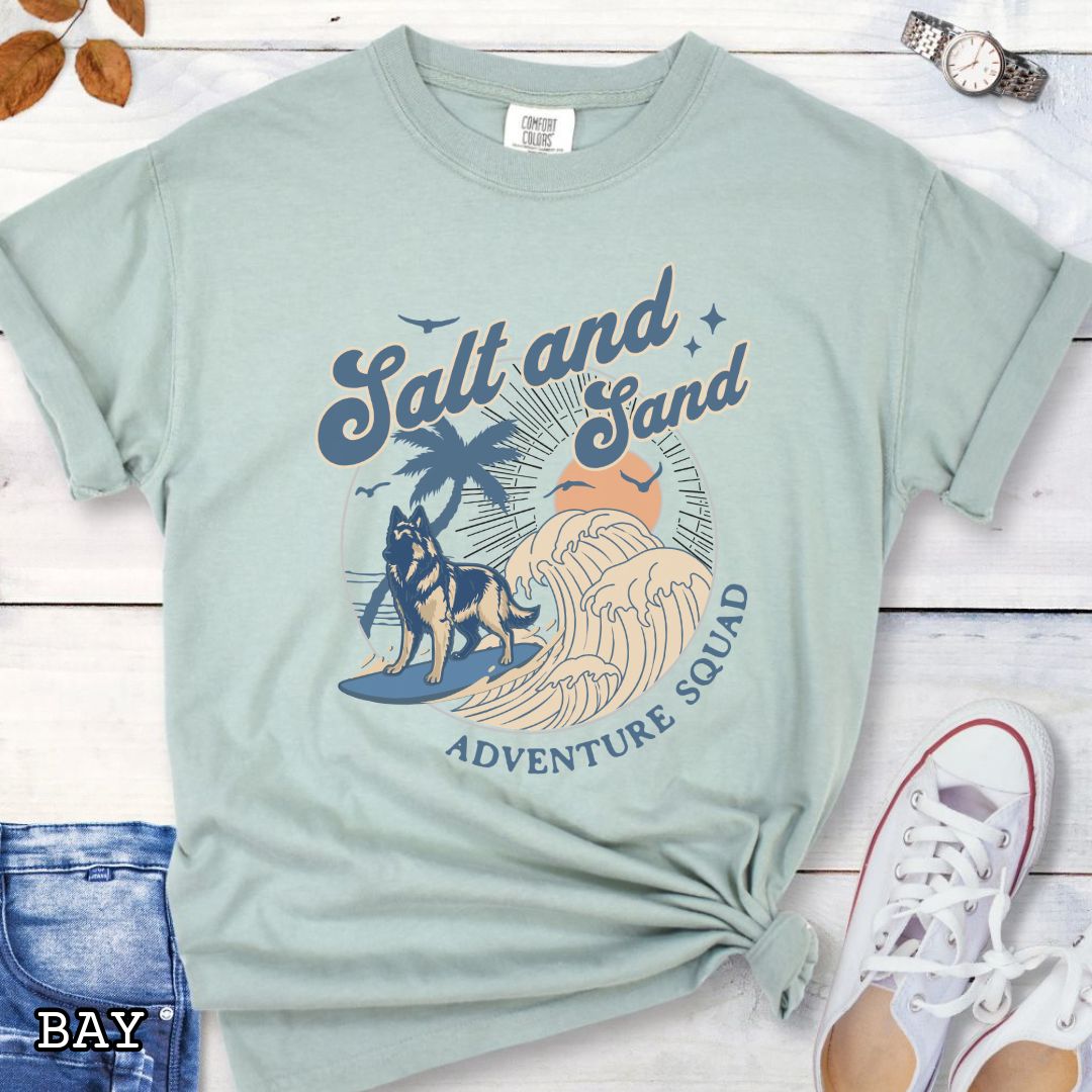 Light green Comfort Colors t-shirt for Belgian Tervuren lovers with beach-themed graphic of a surfing Belgian Tervuren, waves and sunset, and the text 'Salt and Sand Adventure Squad' in retro lettering, shown on a wooden surface with accessories.