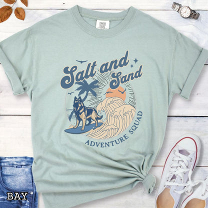 Light green Comfort Colors t-shirt for Belgian Tervuren lovers with beach-themed graphic of a surfing Belgian Tervuren, waves and sunset, and the text 'Salt and Sand Adventure Squad' in retro lettering, shown on a wooden surface with accessories.